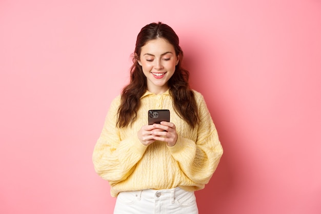 portrait of young brunette woman reading smartphone screen and smiling chatting on social media app shopping online standing against pink wall