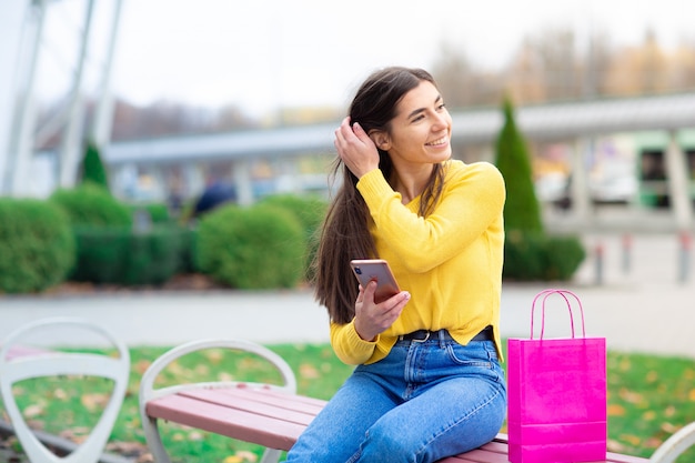 Portrait of young brunette woman sitting outdoor on bench with shopping bags and using mobile phone. Woman dressed in yellow sweater