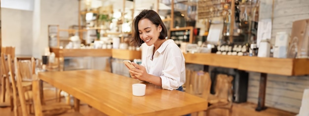 Portrait of young brunette woman sitting with coffee and using smartphone in a cafe chatting on