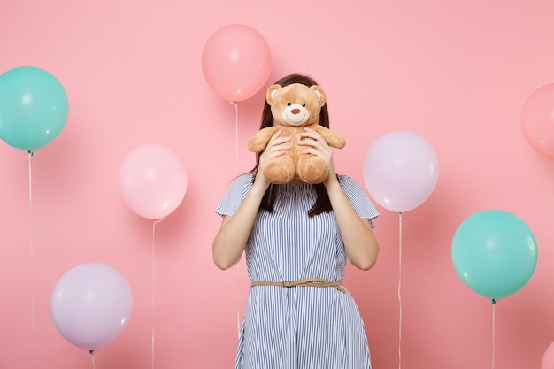 Portrait of young brunette woman wearing blue dress covering face with teddy bear plush toy on pink background with colorful air balloons. Birthday holiday party, people sincere emotions concept.