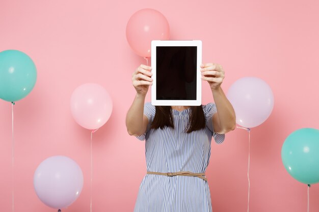 Ritratto di giovane donna castana che indossa vestito blu che tiene il viso coprente con computer tablet pc con schermo vuoto vuoto su sfondo rosa con mongolfiere colorate. concetto di festa di compleanno.