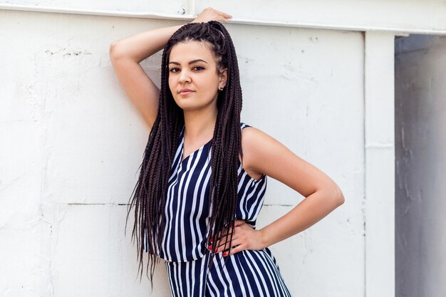 Portrait of young brunette woman with black afro american dreadlocks hairstyle in striped dress standing, posing and looking at camera and smiling. fashion model shot.