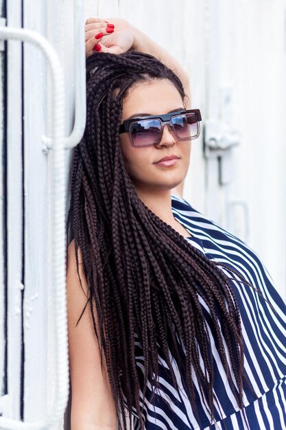 Ritratto di giovane donna bruna con capelli dreadlocks afroamericani neri in abito a righe e occhiali da sole in piedi in posa e guardando la fotocamera in studio indoor girato sul muro bianco