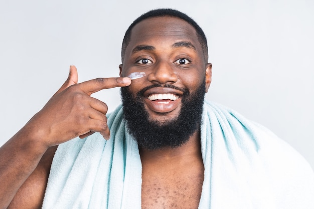 Portrait of young brutal african american black man applying facial cream on his cheek isolated over white background. Close up portrait, men's beauty. skin care