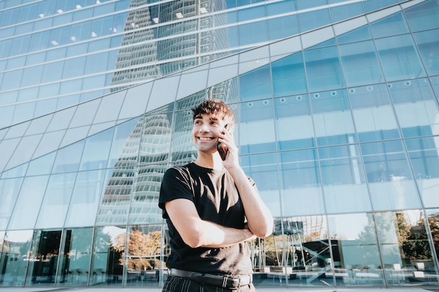 Портрет молодого студента бизнеса, звонящего по телефону, улыбаясь перед массивным зданием с окнами, копией пространства, бизнес-концепция студента ноутбука, бизнес-здания современного дизайна