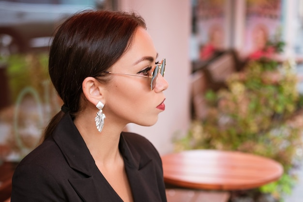 Портрет молодой деловой женщины брюнетки кавказской внешности в черном костюме и сережке сидит ...