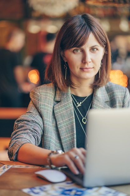 カフェのテーブルに座ってネットブックに取り組んでいる灰色のブレザーの若いビジネス女性の肖像画。コーヒーショップで働くフリーランサー。