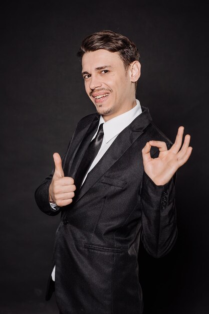 Портрет молодого бизнесмена в черном костюме, показывающего ок, знак, эмоции, выражения лица, чувства, язык тела, знаки, изображение на черном фоне студииxAxAxA