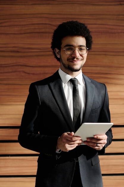 Portrait of young businessman in black suit smiling at camera while using digital tablet standing ag