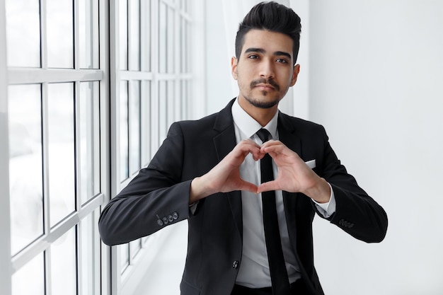 Portrait of young businessman in love shows heart sign against window. service mind business concept