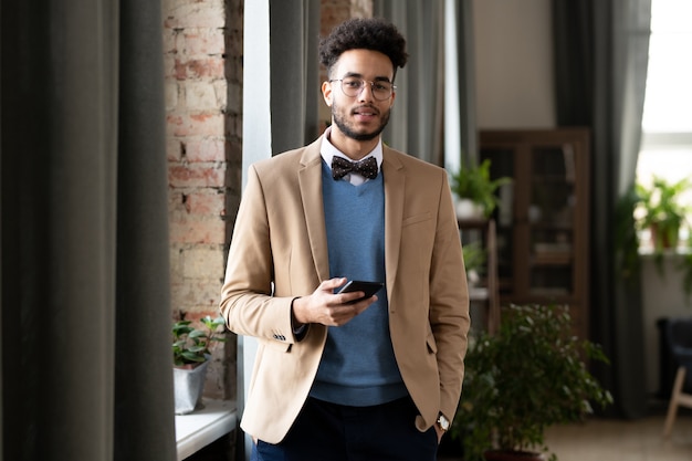 Photo portrait of young businessman in stylish clothing smiling at camera while using his mobile phone at office