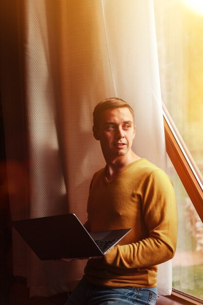 Portrait of young businessman with laptop at window of living room in country house. Workaholic man in home casual clothes working on vacation. Creative inspiration and start-up business. Copy space