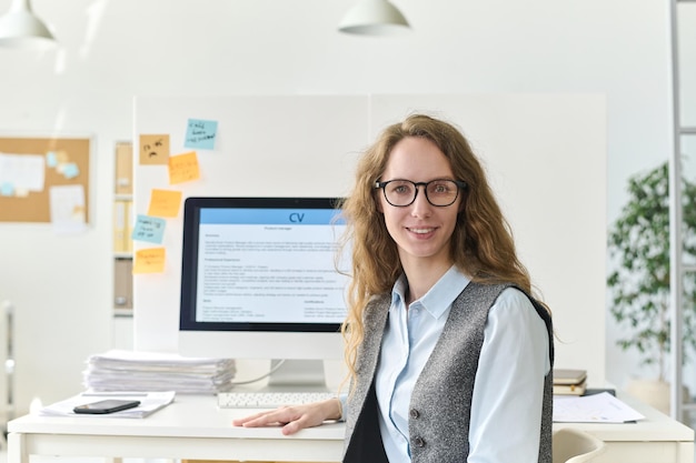 Портрет молодой бизнесменки в очках, улыбающейся в камеру, сидящей на рабочем месте с компьютером