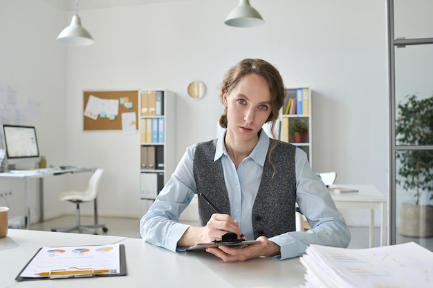 Portrait of young businesswoman looking at camera while making notes in document sitting at her work