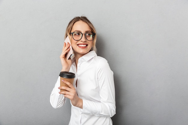 Portrait of young businesswoman wearing glasses holding paper cup of coffee and using cell phone in the office, isolated 