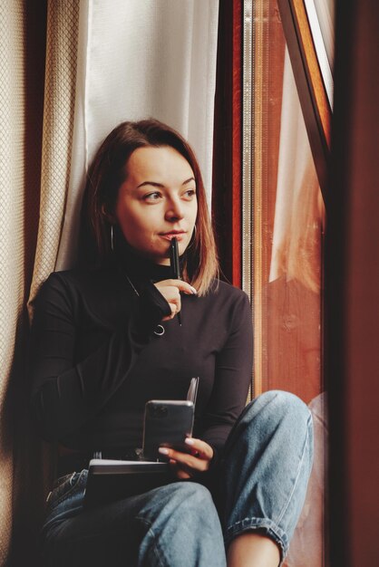 Portrait of young businesswoman with notebook and phone at window of living room in country house. Workaholic woman in home casual clothes working on vacation. Creative inspiration and business