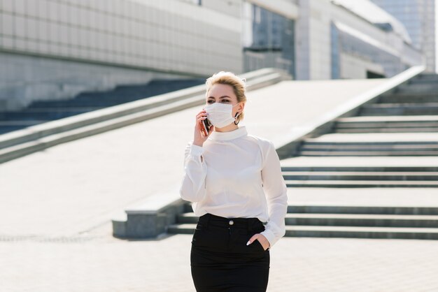 Portrait of a young businesswoman with phone, notebook, tablet, coffee outdoors. Pretty blonde girl in rubber blue gloves and mask.