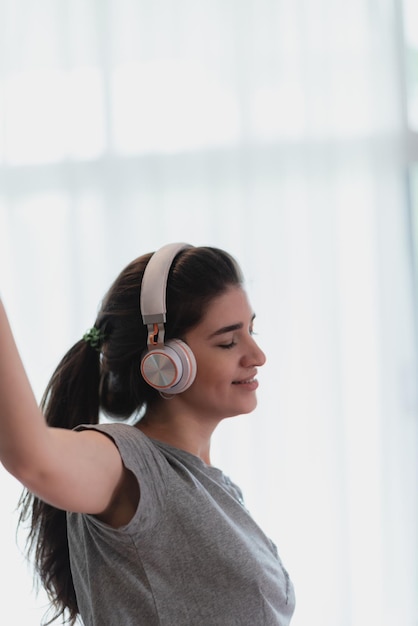 Portrait of young and carefree young woman in casual clothing listening to music using wireless bluetooth headphones dancing and enjoying at home with eyes closed