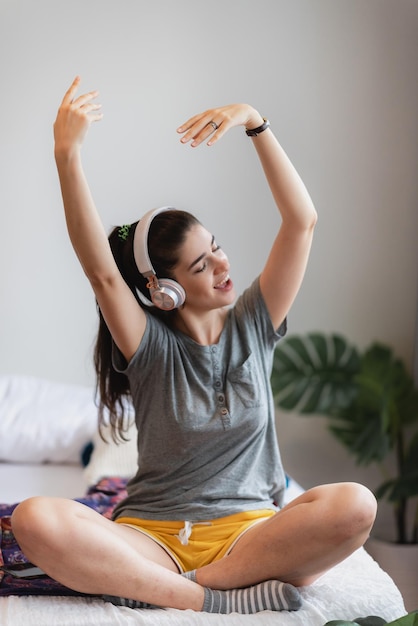 Portrait of young and carefree young woman in casual clothing listening to music using wireless bluetooth headphones dancing and enjoying at home with eyes closed