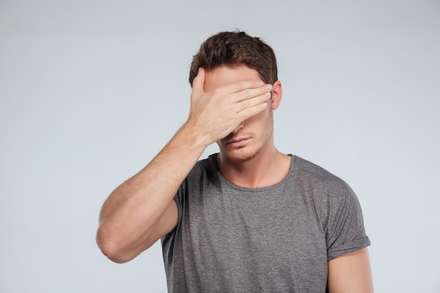 Portrait of a young casual man covering eyes with hand on the gray