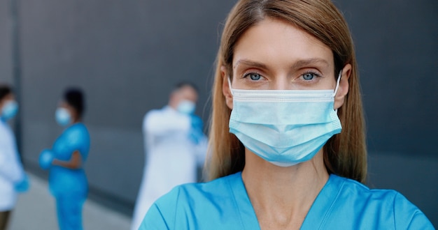 카메라를보고 의료 마스크에 젊은 백인 아름 다운 여자 의사의 초상화. 호흡기 보호에 여성 의사의 닫습니다. 배경에 다중 민족적인 의사 동료입니다.