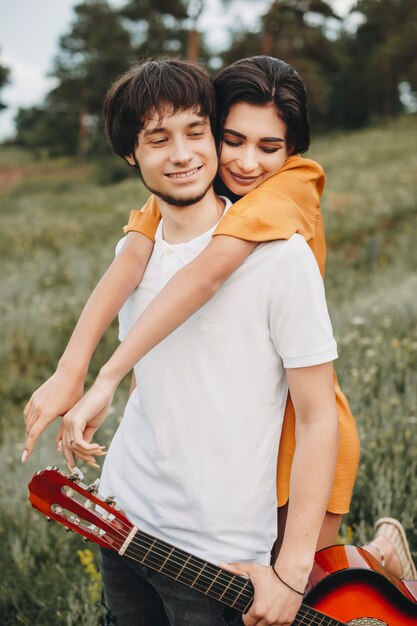 Portrait of a young caucasian couple dating outdoor while man is holding a guitar and girl is embracing him from back.