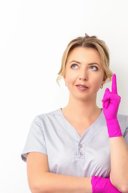 portrait of young caucasian doctor woman wearing rubber gloves in medical uniform with one finger up, looking up against a white background, copy space.