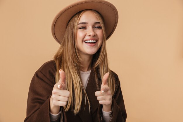 Foto ritratto di giovane ragazza caucasica che indossa cappello e cappotto sorridente e gesticolando fresco isolato su beige