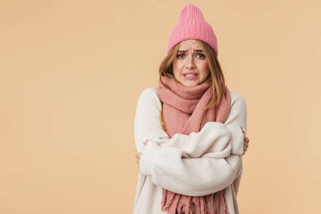 Portrait of young caucasian girl wearing winter hat and scarf trembling and feeling cold isolated on beige