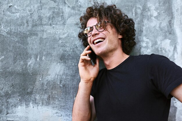 Portrait of young Caucasian happy man standing outdoors and talking on mobile phone Young male with curly hair wears spectacles resting outside making a call on his cell phone on concrete background