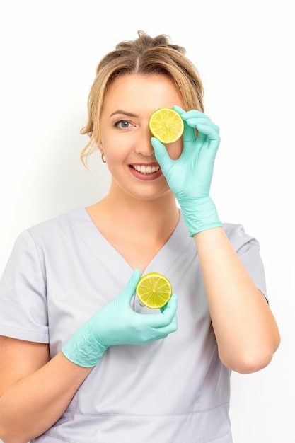 Portrait of young caucasian smiling female beautician covering eye with a lime slice wearing gloves