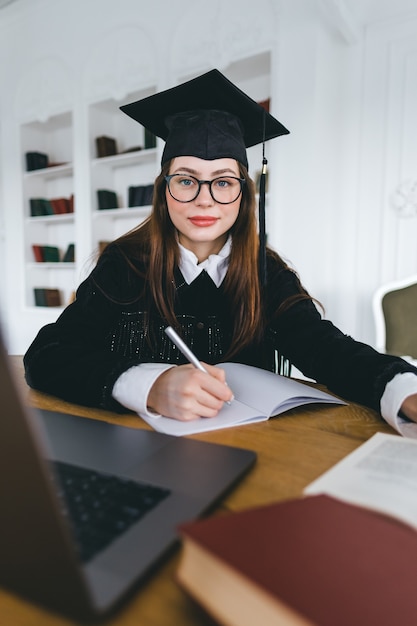 Portrait of young caucasian woman in eyeglasses college student in graduation outfit taking notes