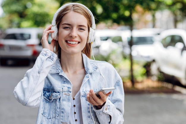 街でスマートフォンとヘッドフォンを使用して幸せに笑っている若い白人女性の肖像画夏の街を歩いている若い女の子