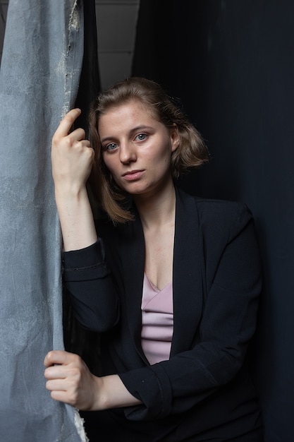 Portrait of young caucasian woman with short hair posing in black suit jacket, holding curtain.