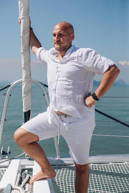 Portrait of a young charismatic man in white clothes standing in a warlike posing on his white yacht and looking into the distance at the floating landscapes. Concept of masculinity