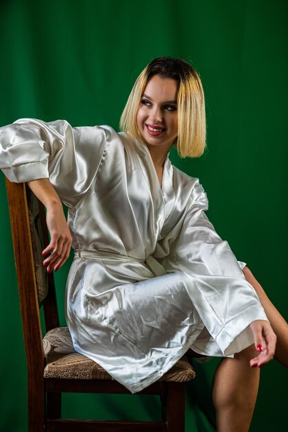 Portrait of a young charming woman in a silk robe sitting on a wooden chair in a studio