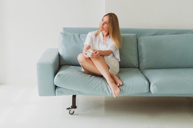 Portrait of a young charming woman in a silk robe with cup of coffee at home on the sofa