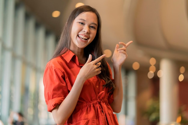 Portrait of young cheerful asian smiling woman hand gesture point to copyspace standing in mall department store with blue bokeh light backgroundsmiling asian female casual cloth smiling look camera