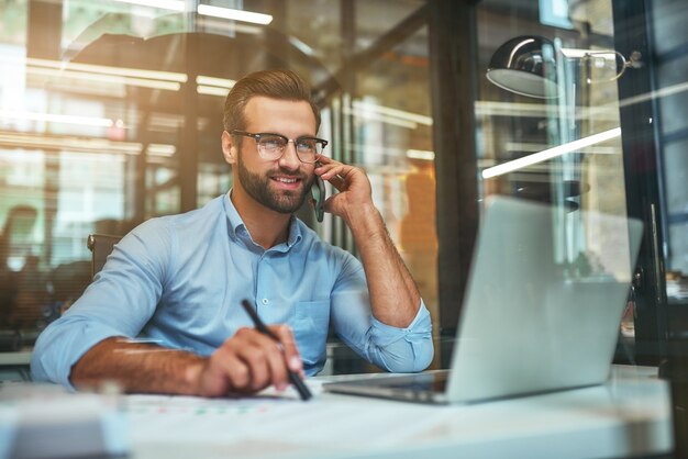 Portrait of young and cheerful businessman in eyeglasses and formal wear talking on the phone