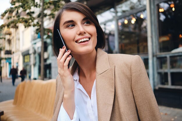 Портрет молодой веселой деловой женщины, счастливо разговаривающей по мобильному телефону в кафе на улице