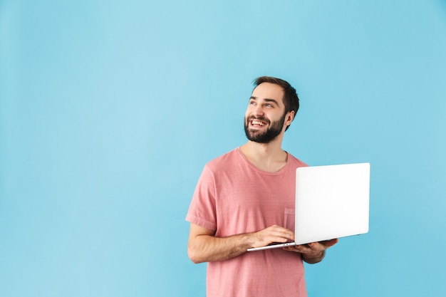 Portrait of a young cheerful excited bearded man wearing t-shirt standing isolated over blue wall, holding laptop computer, looking up