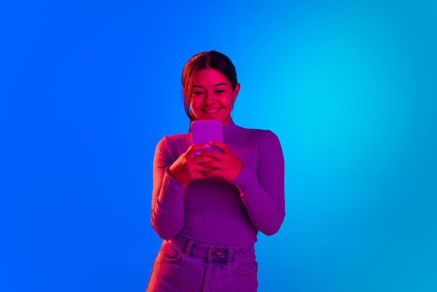 Portrait of young cheerful girl reading text messages on phone smiling isolated over blue background in neon light
