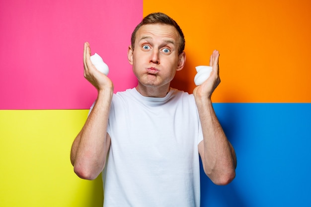 Portrait of a young cheerful guy on a multi-colored background, dressed in a white T-shirt, poured hair foam in his hands, creates a fashionable hairstyle