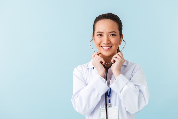 portrait of a young cheerful happy woman doctor posing isolated over blue wall with stethoscope.