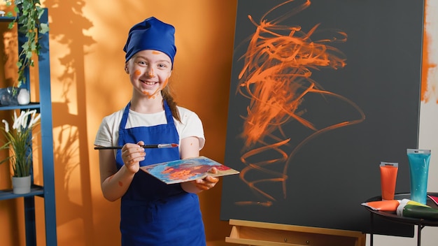 Portrait of young child using orange color paint on canvas, creating art masterpiece with paintbrush and watercolor dye. Little girl making artwork design with aquarelle palette. Handheld shot.