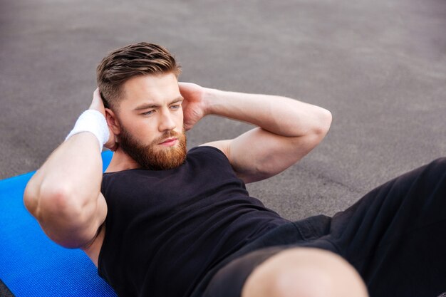 Portrait of a young concentrated handsome sports man doing press workout on fitness mat outdoors