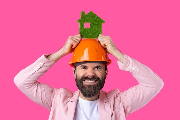 Foto ritratto di giovane ingegnere edile indossare elmetto arancione in una giacca rosa in piedi su sfondo rosso studio un uomo detiene una casa ecologica verde