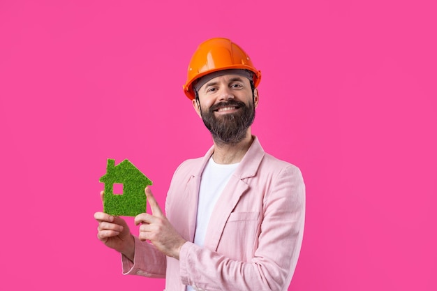 若い建設エンジニアの肖像画は、赤いスタジオの背景に立っているピンクのジャケットにオレンジ色のヘルメットをかぶっています男は緑のエコハウスを保持しています