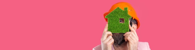 Портрет молодого инженера-строителя в оранжевой каске в розовой куртке, стоящего на красном фоне студии Мужчина держит зеленый экологический дом