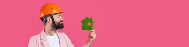 Portrait of young construction engineer wear orange hard hat in a pink jacket standing on red studio background A man holds a green eco house